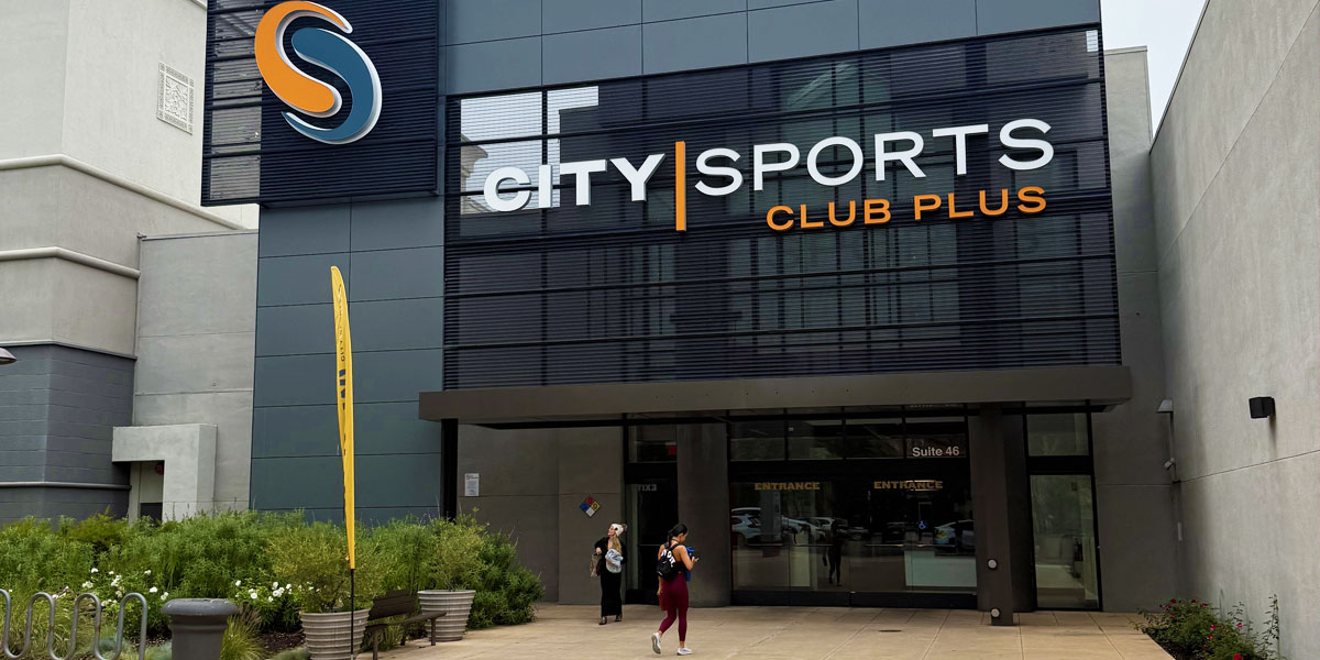 Modern gym entrance with large “City Sports Club Plus” signage on a sleek gray building, glass doors, and people walking near landscaped planters outside.