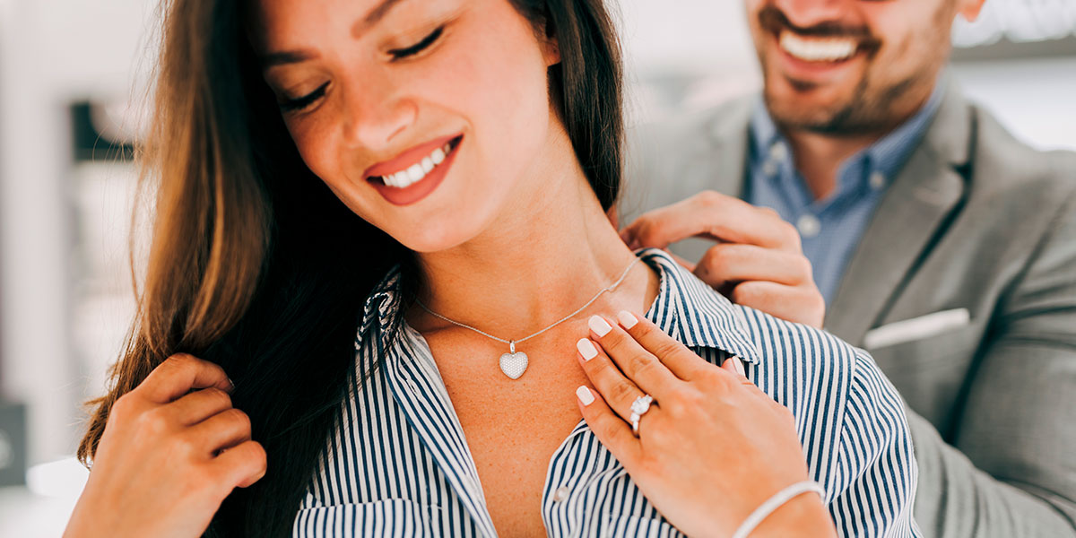 Man standing behind and attractive brunette woman putting a diamond heart necklace around her neck.