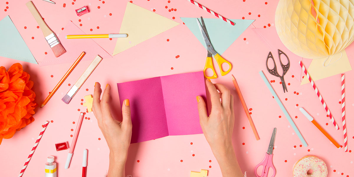 Woman's hands opening a pink card with various art supplies scattered on a pink desk.